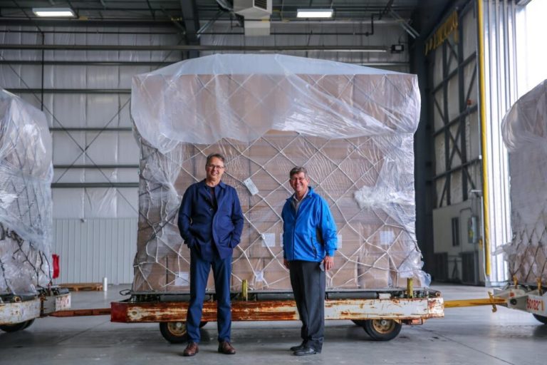 Two men standing in front of a warehouse with boxes covered in plastic.