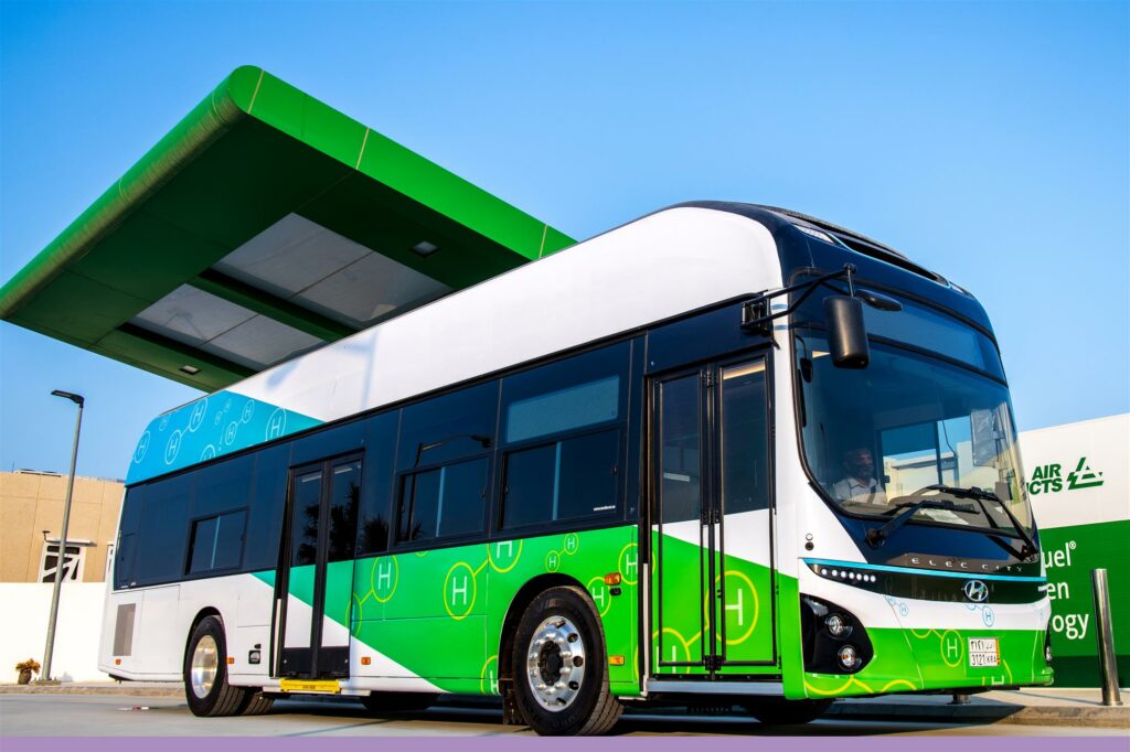 A green and white electric bus parked at a gas station.