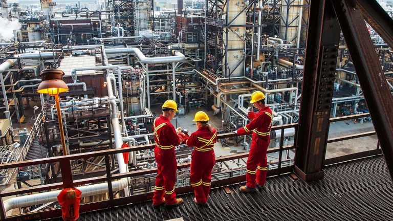 Three workers standing on a railing at an oil refinery.
