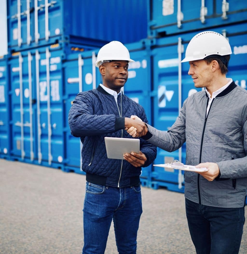 Two men in hard hats shaking hands in front of shipping containers.