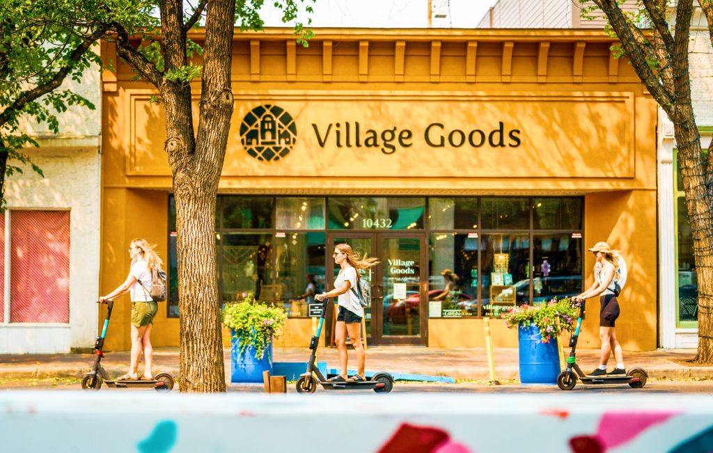A group of people riding scooters in front of a store called village goods.